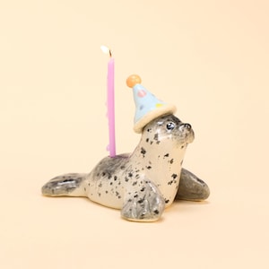 May include: A ceramic seal figurine with a light blue party hat and a lit pink candle. The seal is white with black spots and has a playful expression. The candle is tall and slender, adding a festive touch to the design.