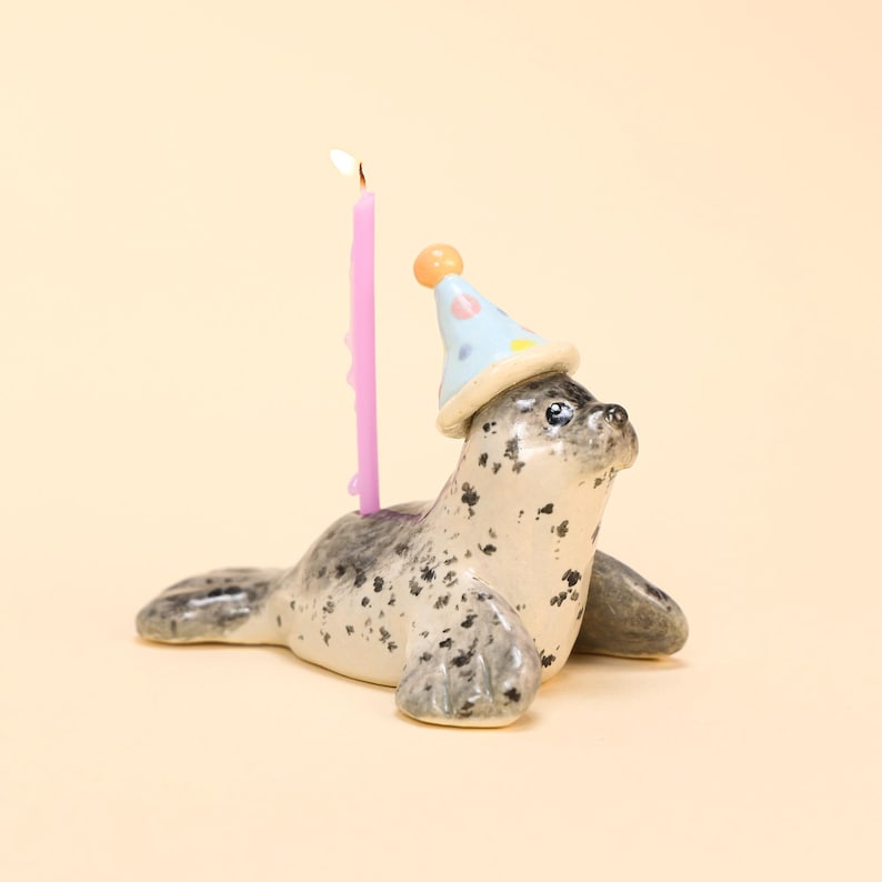 May include: A ceramic seal figurine with a light blue party hat and a lit pink candle. The seal is white with black spots and has a playful expression. The candle is tall and slender, adding a festive touch to the design.
