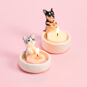 May include: Two ceramic candle holders, one with a cat and the other with a dog, each holding a lit candle. The cat candle holder has a pink candle, while the dog has a white candle. The background is a solid pink.