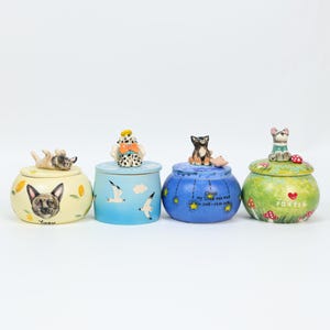 May include: Four decorative ceramic jars with lids, each featuring a different dog or cat figurine on top. The jars are painted in various colors, including yellow, blue, and green, with whimsical designs and text.