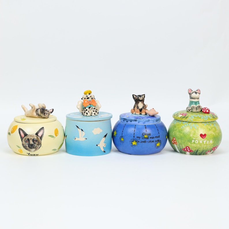 May include: Four decorative ceramic jars with lids, each featuring a different dog or cat figurine on top. The jars are painted in various colors, including yellow, blue, and green, with whimsical designs and text.