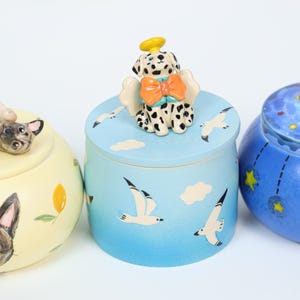 May include: A collection of ceramic jars with lids, each featuring a unique animal-themed design. One jar has a Dalmatian dog with angel wings and a bow, another has a dog, and the third has a celestial design with stars.