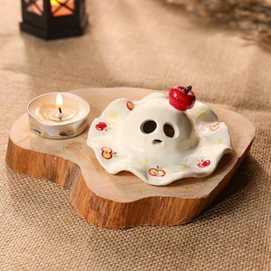 May include: A white ceramic ghost-shaped candle holder with a red apple on top, decorated with red apples and yellow stars. A lit tealight candle sits on a wooden base. The background includes a small black lantern.
