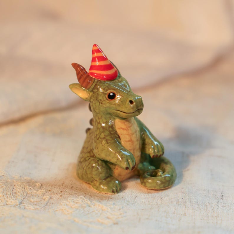 K&ouml;nnte beinhalten: Eine Keramikfigur eines gr&uuml;nen Drachen mit einem rot-orange gestreiften Partyhut. Der Drache hat einen hellgr&uuml;nen K&ouml;rper, einen beigen Bauch und braune H&ouml;rner. Der Drache sitzt mit eingerolltem Schwanz.