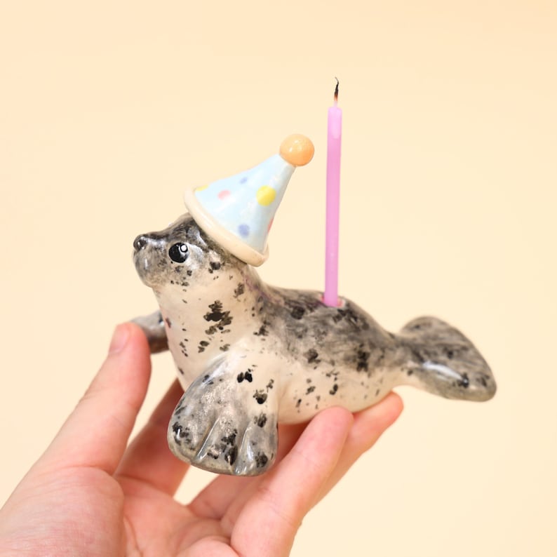 May include: A ceramic seal figurine with a party hat and a pink candle. The seal is gray and white with black spots. The party hat is light blue with colorful polka dots. The candle is lit.