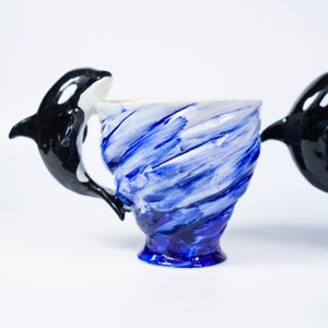 May include: A blue and white ceramic mug with a black and white orca whale handle. The mug is shaped like a wave and has a blue and white swirl design.