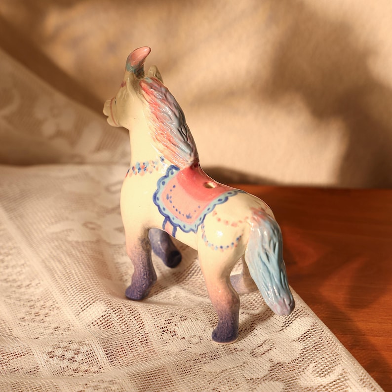 Op de afbeelding: Een keramieken paardenbeeldje met een cr&egrave;mekleurig lichaam, versierd met roze, blauwe en paarse accenten. De manen en staart hebben een mix van roze en blauwe tinten. Het paard heeft een decoratief zadel en staat op een wit kanten oppervlak.