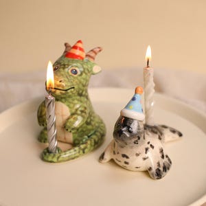 Pode incluir: Uma estatueta de dragão de cerâmica verde com um chapéu de festa às riscas vermelhas e amarelas e uma estatueta de foca branca e cinzenta com um chapéu de festa azul com pintas cor de laranja. Cada figura segura uma vela acesa.