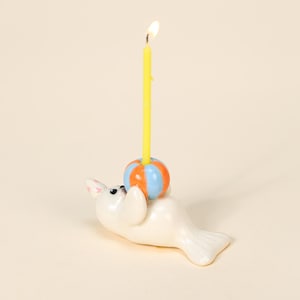 Può includere: Una statuetta di foca in ceramica bianca che tiene una palla a strisce con una candela arancione, blu e gialla. La candela è accesa, con una piccola fiamma in cima. La foca è sdraiata, con la testa e le pinne sollevate.