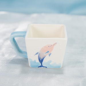 Op de afbeelding: Een vierkante witte keramische mok met een lichtblauw handvat. De mok heeft een aquarel illustratie van een dolfijn die uit blauwe golven springt, met roze en blauwe kleuren. De mok heeft een lichtblauw handvat.