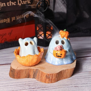 May include: Two ceramic Halloween ghost figurines. One ghost is inside an orange pumpkin, and the other is holding a jack-o'-lantern basket with a pink candy. Both ghosts are light blue with black eye holes.