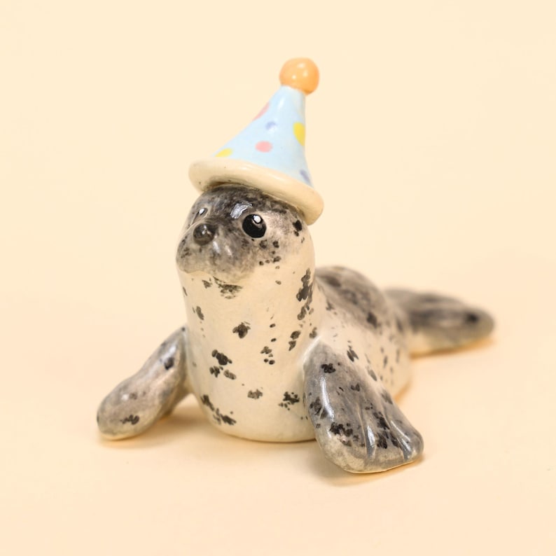 May include: A ceramic seal figurine with a party hat. The seal is primarily white with black spots and gray flippers. The party hat is light blue with yellow hearts and pink polka dots, topped with an orange sphere.
