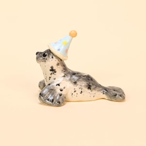 May include: A ceramic seal figurine with a light blue party hat adorned with colorful polka dots. The seal has a speckled gray and white body, with a playful expression. The figurine is set against a soft, neutral background.
