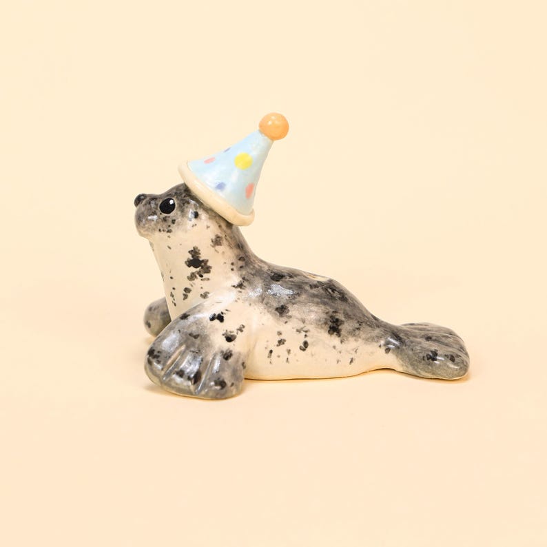 May include: A ceramic seal figurine with a light blue party hat adorned with colorful polka dots. The seal has a speckled gray and white body, with a playful expression. The figurine is set against a soft, neutral background.