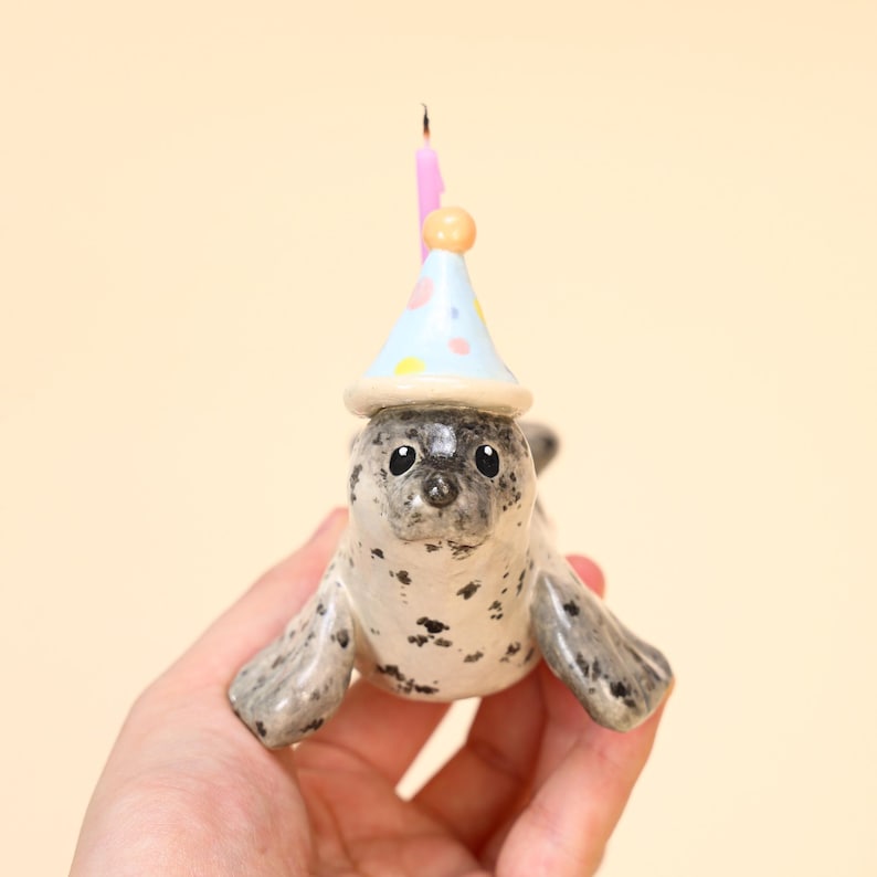 May include: A ceramic seal figurine with a party hat and a lit pink candle. The seal is gray and white with black spots. The party hat is light blue with colorful polka dots. The candle has an orange ball on top.