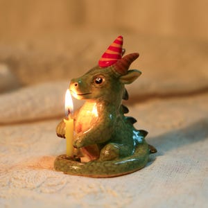 K&ouml;nnte beinhalten: Eine gr&uuml;ne Keramik-Drachenfigur, die eine brennende gelbe Kerze h&auml;lt. Der Drache tr&auml;gt einen rot-orange gestreiften Partyhut. Die Figur ist ein dekorativer Gegenstand. Die Kerze ist etwa 10 cm hoch.
