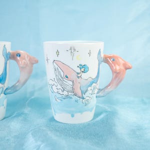 Puede incluir: Una taza de cer&aacute;mica blanca con una ballena azul y una peque&ntilde;a persona montando en su espalda. La taza tiene un asa de delf&iacute;n rosa.