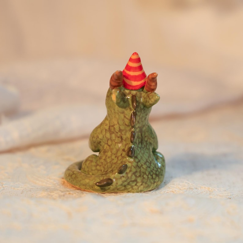 K&ouml;nnte beinhalten: Keramikfigur eines gr&uuml;nen Drachen mit strukturierter, schuppiger Erscheinung. Der Drache tr&auml;gt einen Partyhut mit roten und orangefarbenen Streifen. Die Figur hat braune H&ouml;rner und einen braunen R&uuml;cken.