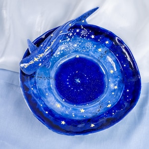 May include: A blue ceramic dish with a white whale design and gold stars. The dish is decorated with a galaxy-like pattern.