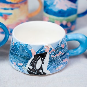 Op de afbeelding: Een blauwe en witte keramische mok met een handgeschilderd ontwerp van een gestileerde walvis en oceaan golven. De mok heeft een wit interieur en een blauwe handgreep.
