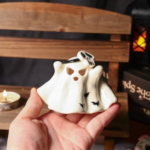 Könnte beinhalten: Ein weißer Keramik-Geist-Kerzenhalter mit Schädel- und Fledermaus-Design. Der Geist hat ausgeschnittene Augen und Mund, und schwarze Fledermäuse sind auf der Unterseite bemalt.