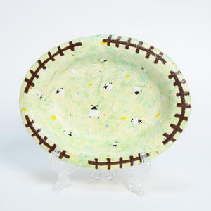 Puede incluir: Plato ovalado de cerámica con fondo verde claro y un patrón de ovejas. El borde presenta un diseño cosido marrón. El plato se exhibe sobre un soporte de plástico transparente.