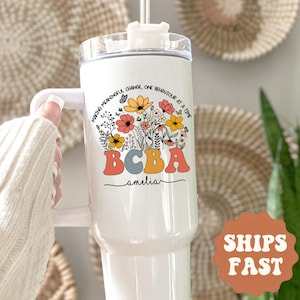 May include: White insulated tumbler with a handle, featuring a floral design with the text "MAKING MEANINGFUL CHANGE, ONE BEHAVIOUR AT A TIME" and "BCBA amelia" in colorful letters. The tumbler has a white lid and a straw.