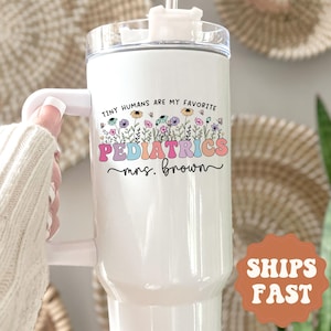 May include: White insulated tumbler with a handle and a clear lid. The tumbler features the text "TINY HUMANS ARE MY FAVORITE PEDIATRICS mrs. brown" in colorful lettering with floral accents. The tumbler is designed for beverages.