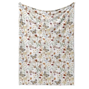 a white blanket with a floral print pattern, including butterflies and flowers. The blanket has the name "Riley" written on it in a cursive font.