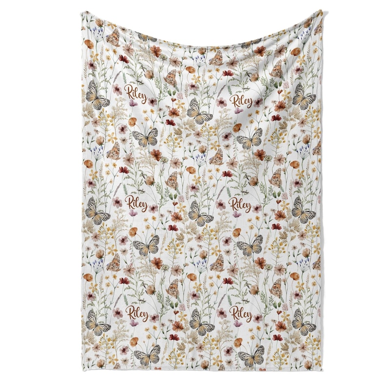 a white blanket with a floral print pattern, including butterflies and flowers. The blanket has the name "Riley" written on it in a cursive font.