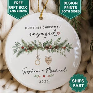 May include: A round white ceramic ornament featuring the text OUR FIRST CHRISTMAS engaged with a small red heart. Below, a green pine branch illustration with pinecones and berries hangs a gold ring and a heart-shaped tag reading forever. Sophia + Michael 2026.
