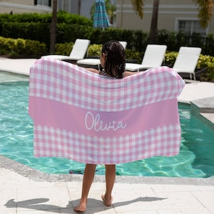 May include: A pink and white gingham beach towel with the name "Olivia" printed in white on a pink stripe.
