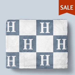 May include: A soft, white and blue blanket with a checkerboard pattern. Each blue square features a large white letter "H". The blanket is folded and displayed against a light blue background. A red "SALE" tag is in the upper right corner.