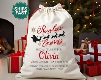 May include: A cream-colored Christmas gift bag with a red drawstring. The bag features the text "Reindeer Express" and "Special Delivery For Olivia" in red. A black silhouette of Santa's sleigh and reindeer is also visible. The bag is surrounded by wrapped gifts.