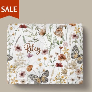 a white blanket with a floral and butterfly pattern. The blanket has the name "Riley" written on it in cursive.