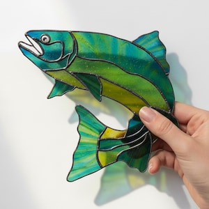 Può includere: Un pesce in vetro colorato nei toni del verde, blu e giallo. Il pesce sta saltando, con la bocca aperta e le pinne spiegate. L'opera d'arte ha contorni neri ed è tenuta su uno sfondo bianco.