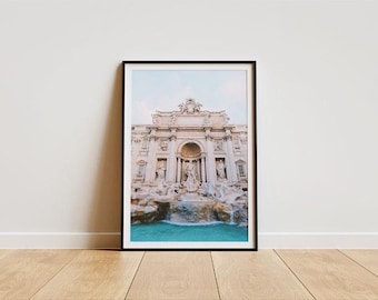 May include: A framed print of the Trevi Fountain in Rome, Italy. The photograph captures the baroque architecture, cascading water, and turquoise pool. The print is in a black frame, set against a white wall and wooden floor.