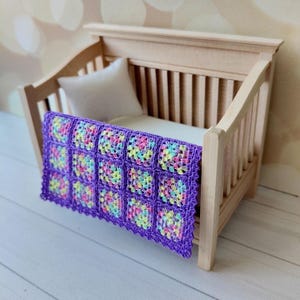 Puede incluir: Una cuna de madera en miniatura con una almohada blanca y una vibrante manta tejida a mano. La manta presenta un borde morado y cuadrados coloridos en tonos rosa, amarillo y azul. La cuna es de madera clara.