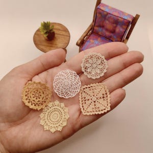 May include: Five miniature crochet doilies in various shades of beige, cream, and white. The collection includes four circular doilies with intricate floral and spiral patterns and one square doily with a geometric openwork design.