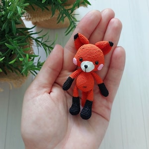 May include: A small, handmade orange fox amigurumi with black detailing on the ears, feet, and arms. The fox has a white muzzle, pink cheeks, and black eyes. The crochet fox is held in a hand.