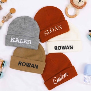 Peut inclure: Quatre bonnets en tricot de différentes couleurs avec des noms personnalisés : KALEO, SLOAN, ROWAN et CUSTOM. Les bonnets sont disposés sur une surface blanche.
