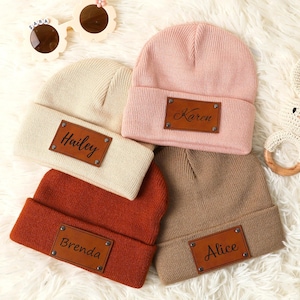 May include: Four knitted beanies in different colours with personalised leather patches. The beanies are beige, pink, brown, and orange. The patches have the names Hailey, Karen, Alice, and Brenda.