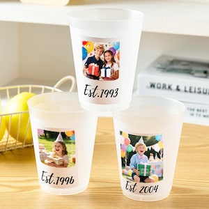 以下が含まれることがあります： 半透明のプラスチック製カップ3個。それぞれにパーソナライズされた写真と黒い文字が印刷されています。上のカップには子供2人の写真と「Est. 1993」の文字、左下のカップには子供と「Est. 1996」、右下のカップには子供と「Est. 2004」の文字が印刷されています。