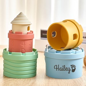 Puede incluir: Juego de juguetes apilables con diseño de torre con techo beige, sección media coral con números y base verde. Un cilindro amarillo se encuentra sobre un recipiente azul claro con el nombre "Hailey" y una huella. Juguete educativo.