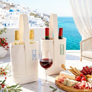 May include: Two off-white canvas wine tote bags with single handles, each featuring a vertical embroidered monogram letter in blue or green. The bags hold wine bottles and sit on a table with a glass of red wine and a wooden charcuterie board.