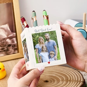 Puede incluir: Un portalápices blanco estilo bloque con fotos familiares y el texto "Best Dad Ever!" es sostenido en las manos de una persona. El portalápices contiene tres bolígrafos con detalles rojos, verdes y dorados. Un patito de goma amarillo y un tren de madera están en el fondo.