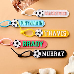 May include: A collection of personalized soccer-themed keychains in various colors. Each keychain features a soccer ball design and a name in bold letters. The keychains are in white, blue, yellow, red, and black.