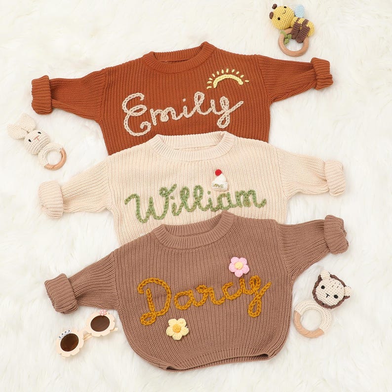 Op de afbeelding: Drie gebreide truien met geborduurde namen: "Emily", "William" en "Darcy". Elke trui heeft een uniek ontwerpelement: een zon, een klein taartje en een bloem.
