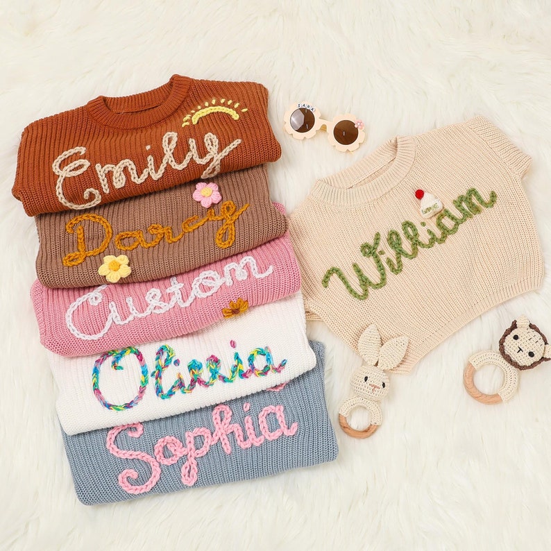 Op de afbeelding: Een stapel van vijf gebreide truien met verschillende namen geborduurd in kleurrijk garen. De namen zijn Emily, Darcy, Custom, Olivia en Sophia. De truien zijn in verschillende kleuren, waaronder bruin, roze, wit en blauw.