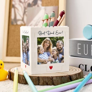 May include: A white block-style pen holder with the text "Best Dad Ever!" and a family photo. The holder has a red heart and holds pens. It sits on a wooden slice with a yellow rubber duck and pencils nearby.